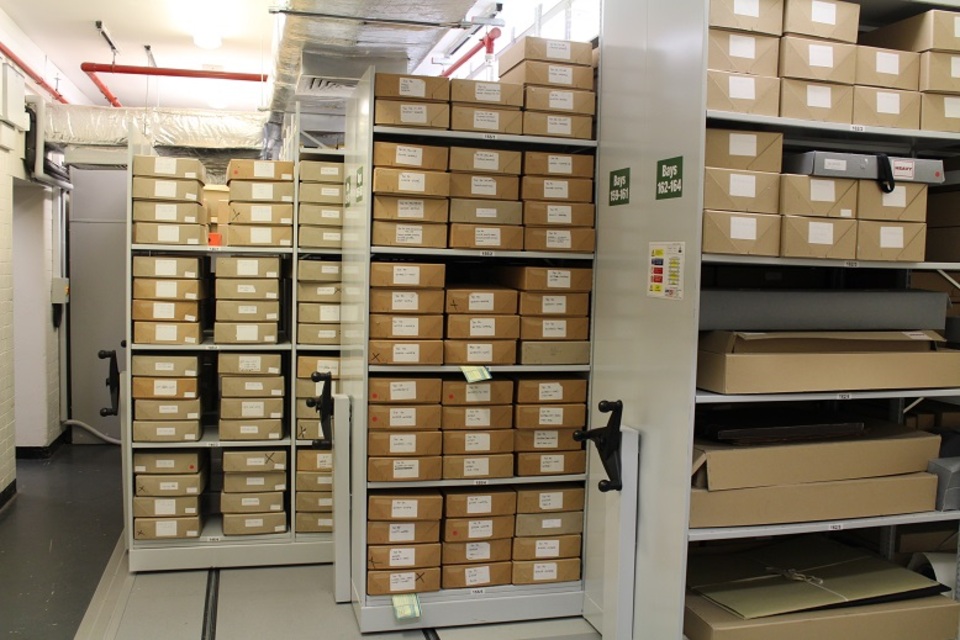 Archive storage room with shelves of brown cardboard boxes, each labeled with white tags. The shelves are metal and movable, with handles for sliding.